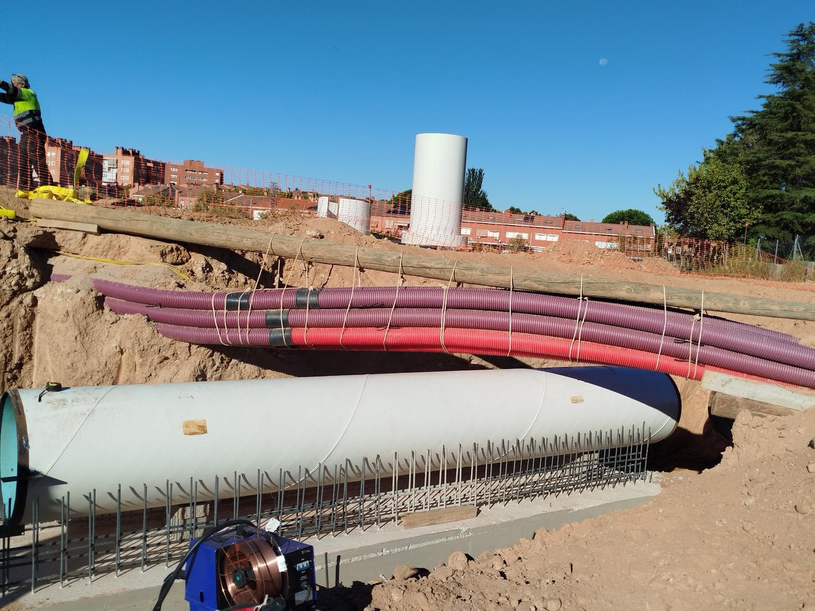 Tubería de acero de gran diámetro montada sobre su cama de hormigón armado en la zanja, con las canalizaciones de servicios cruzando por encima.